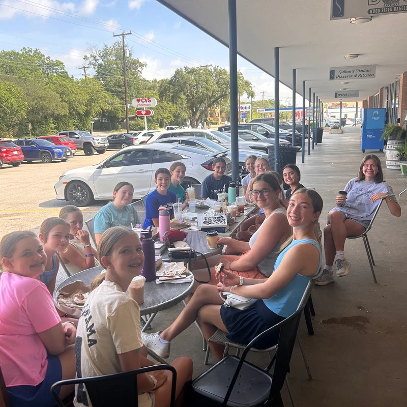 A large group of teenage girls smiling while sitting at tables on a covered patio outside a bagel shop, enjoying food and drinks on a sunny day.