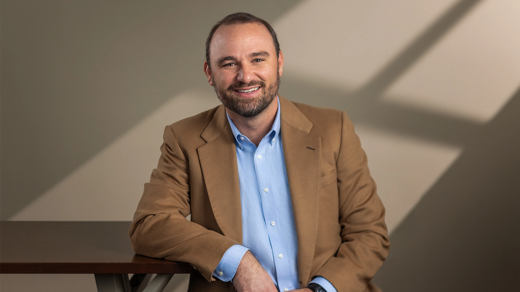 A smiling man with a beard, wearing a blue shirt and tan blazer, leans on a dark table in a studio setting with dramatic shadows on the wall behind him.