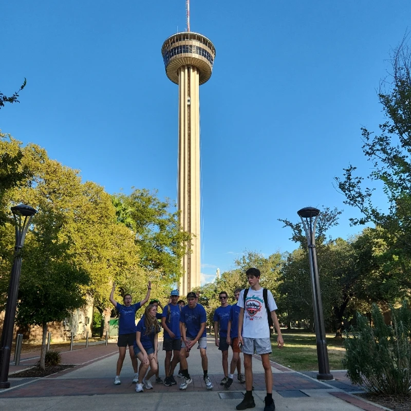 A group of young people pose playfully on a park walkway, with the tall Tower of the Americas rising behind them against a clear blue sky.