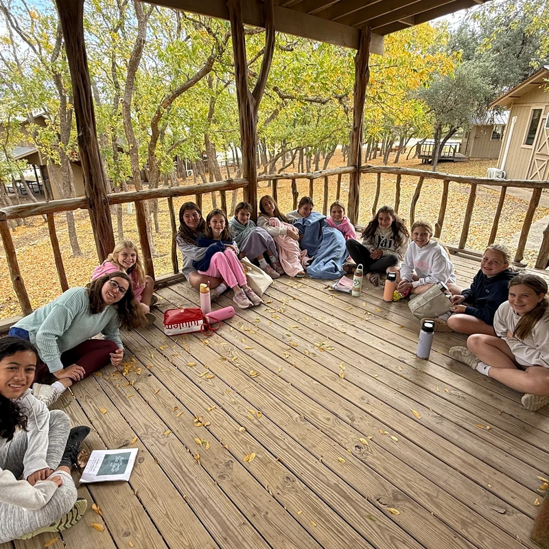 A group of young girls smiles while sitting together on a rustic wooden porch, surrounded by cabins and trees with fall foliage.