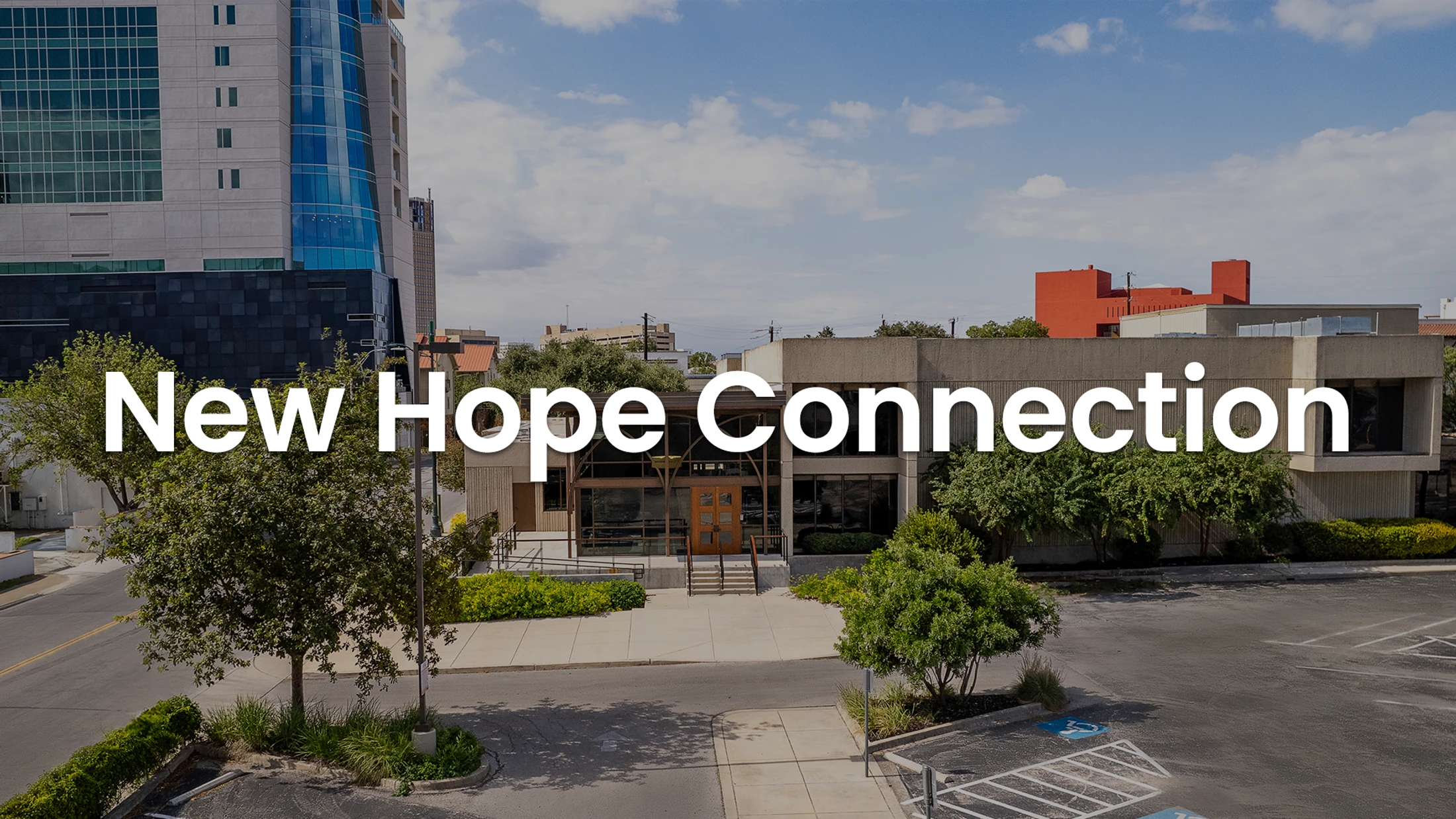 The exterior of the New Hope Connection building, a modern structure with a parking lot, viewed from a high angle against a city skyline on a sunny day.