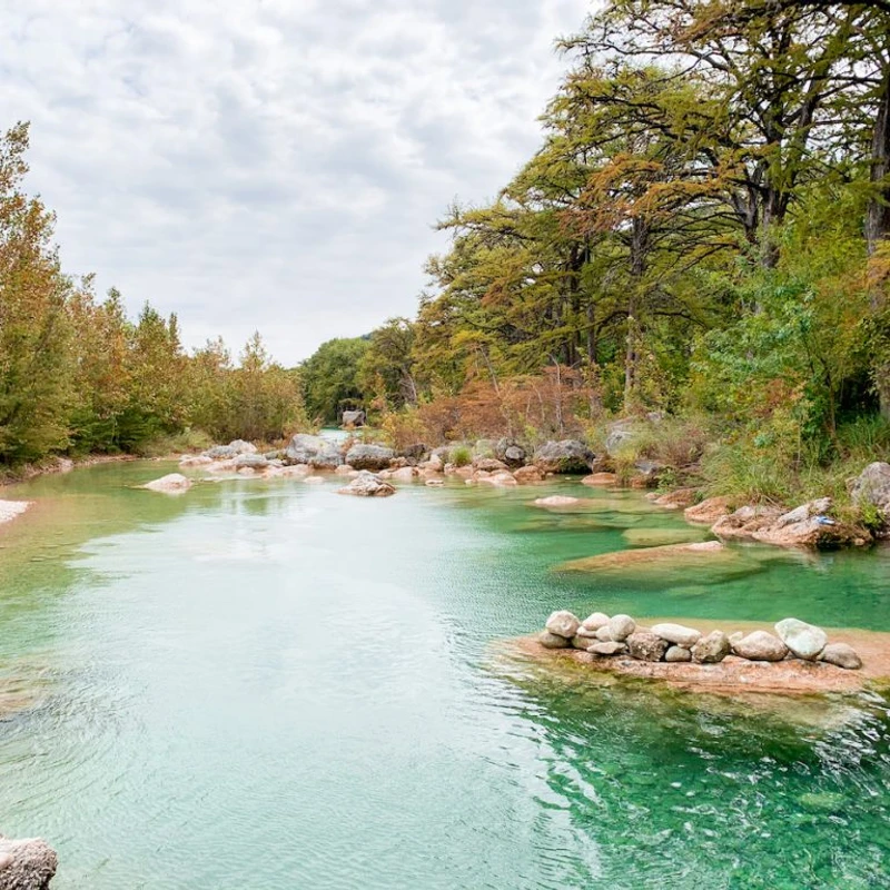 A clear, turquoise river flows gently through a forest with trees showing autumn colors. Rocks are visible in the water and along the banks under a cloudy sky.
