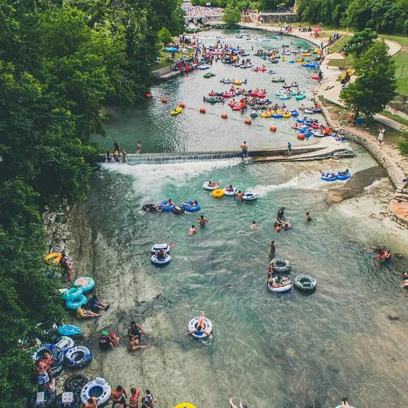 A high-angle shot of a wide river filled with hundreds of people floating in colorful inner tubes, surrounded by lush green trees on a summer day.