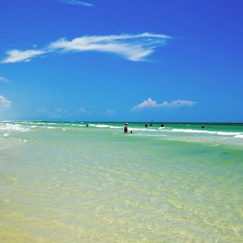 A wide beach scene on a sunny day with people swimming and relaxing in clear, shallow turquoise water under a vast blue sky with a few clouds.