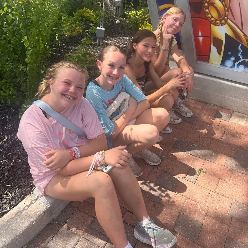 Four young girls sit in a single-file line on a brick sidewalk in the sunshine, smiling at the camera. They are wearing casual summer clothes.