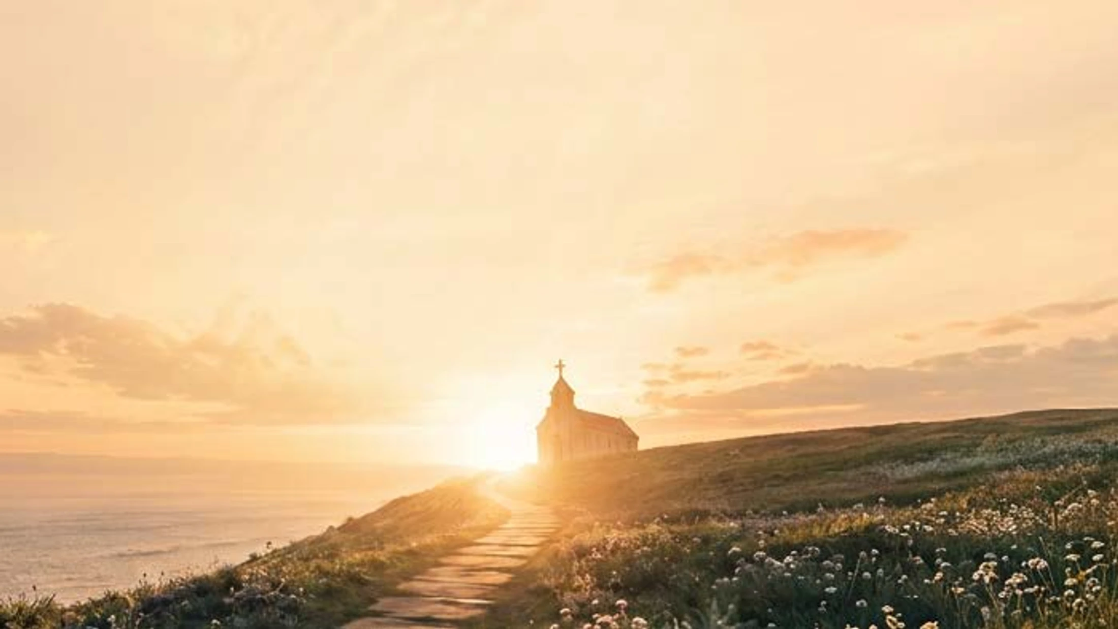 A stone path winds up a grassy hill toward a small church, silhouetted against a golden sunset over the ocean. Wildflowers dot the hillside.