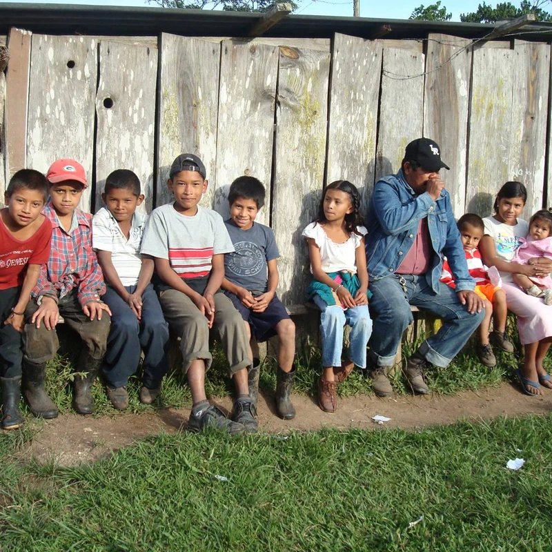 A group of nine children and one man sitting in a row on a wooden bench in front of a weathered wooden building in a grassy, rural area.