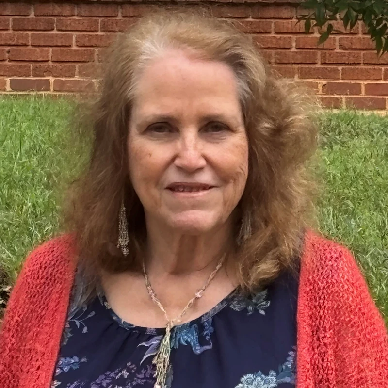 A headshot of a smiling woman with light brown, shoulder-length hair. She wears a red cardigan and stands outdoors in front of a brick wall and grass.