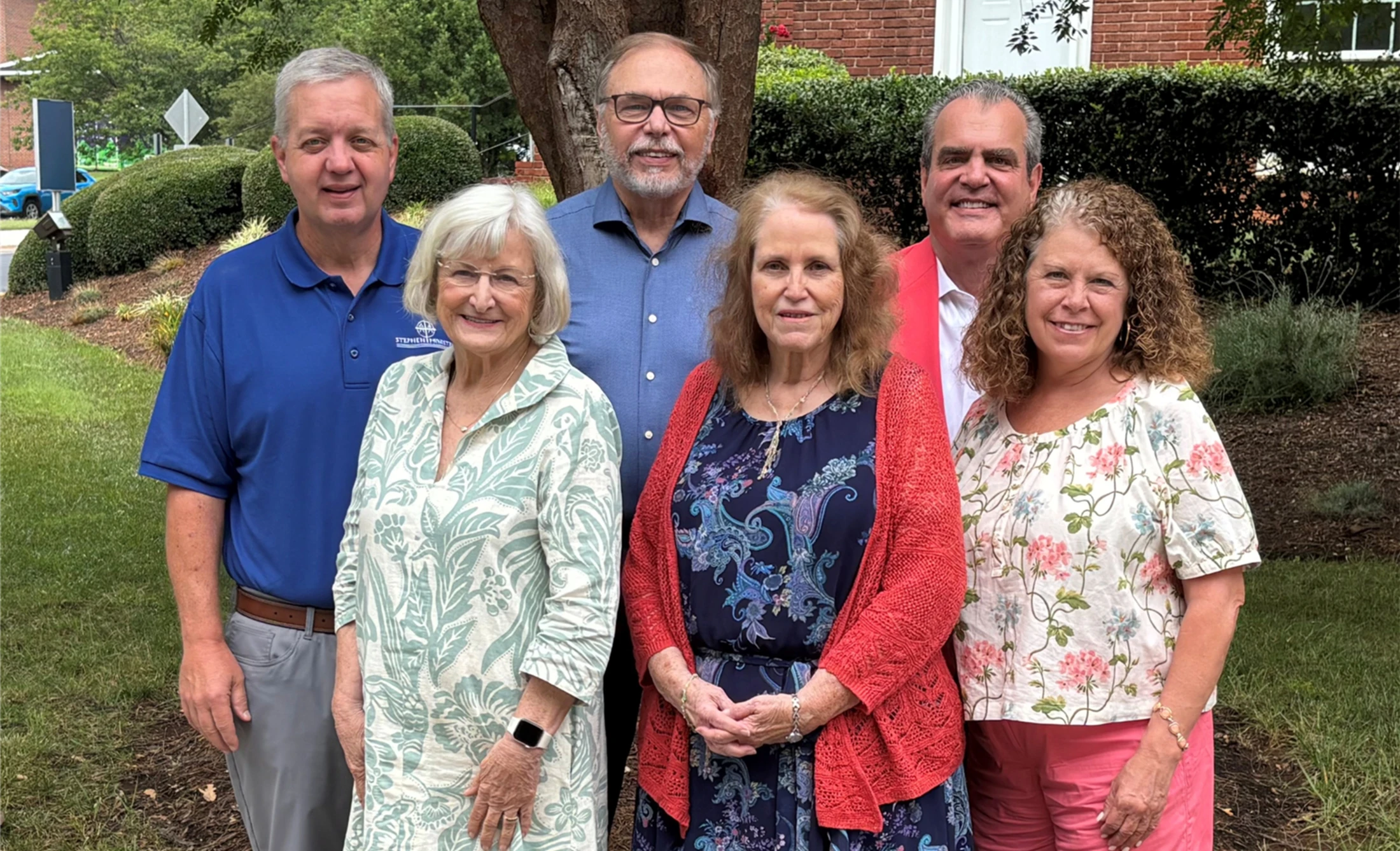 A group of six smiling adults, three men and three women, stand together for a photo outdoors in a grassy area with trees and a brick building behind them.