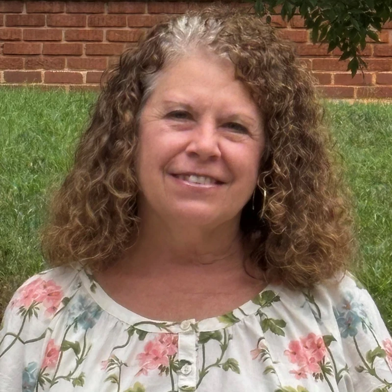 A headshot of a smiling middle-aged woman with curly brown and gray hair. She is wearing a floral blouse and stands outside in front of a brick wall and grass.