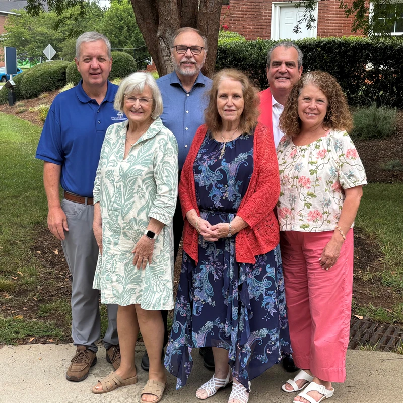 A group of six smiling adults, three men and three women, stand together for a photo outdoors in front of a tree, bushes, and a brick building.