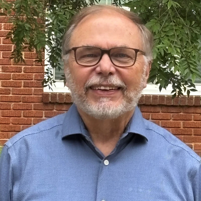 A head-and-shoulders portrait of a smiling older man with a grey beard and glasses. He wears a blue shirt and stands in front of a brick wall and green leaves.