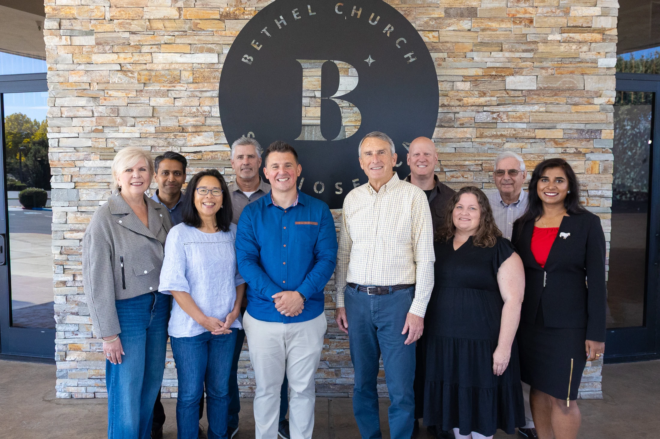 A diverse group of ten men and women stand smiling in front of a stone wall with a large circular sign for Bethel Church San Jose.