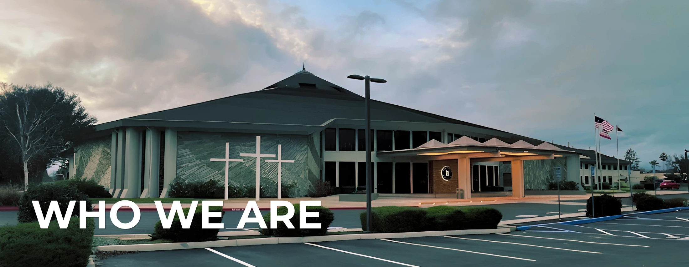 A modern church building at dusk with three white crosses on the wall and an empty parking lot. The text 'WHO WE ARE' is in the foreground.