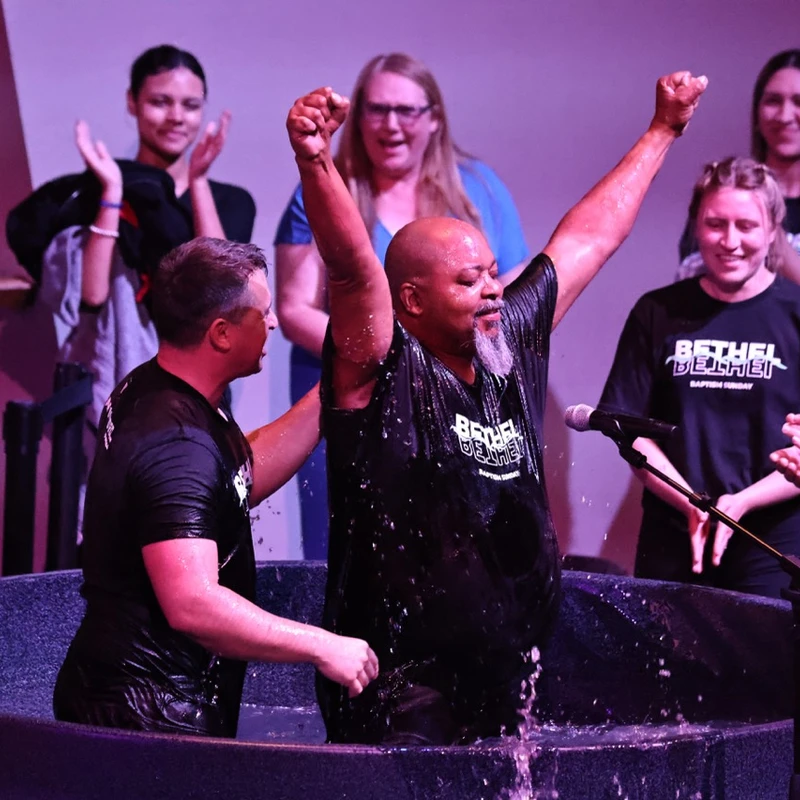 A man with arms raised in triumph emerges from the water of a baptismal font, as another man assists and a congregation watches and claps.