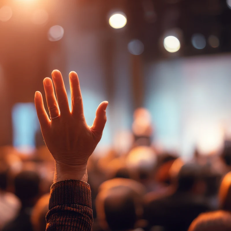 A person raises their hand in a crowded auditorium during a conference. The focus is on the hand, with the audience and stage blurred in the background.