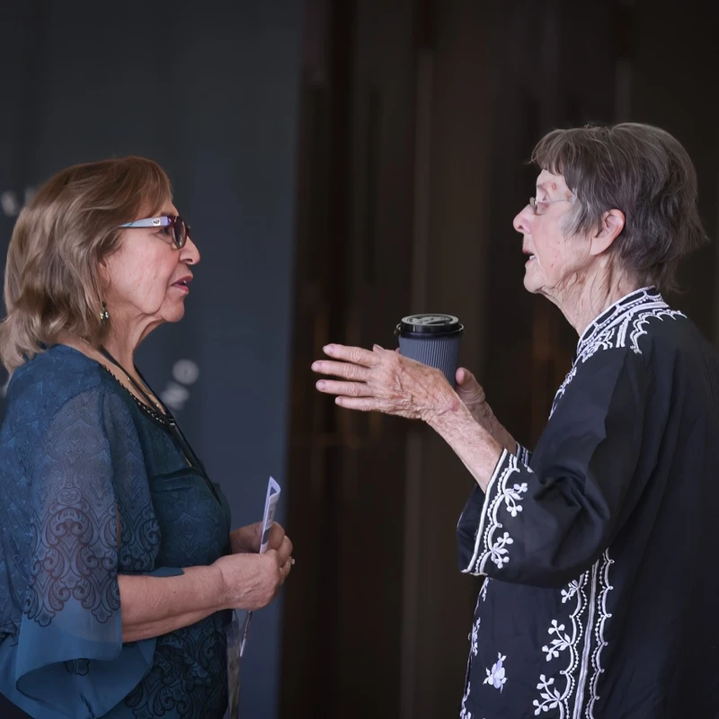 Two older women stand facing each other, engaged in conversation. One woman gestures with her hands while holding a coffee cup, and the other listens intently.
