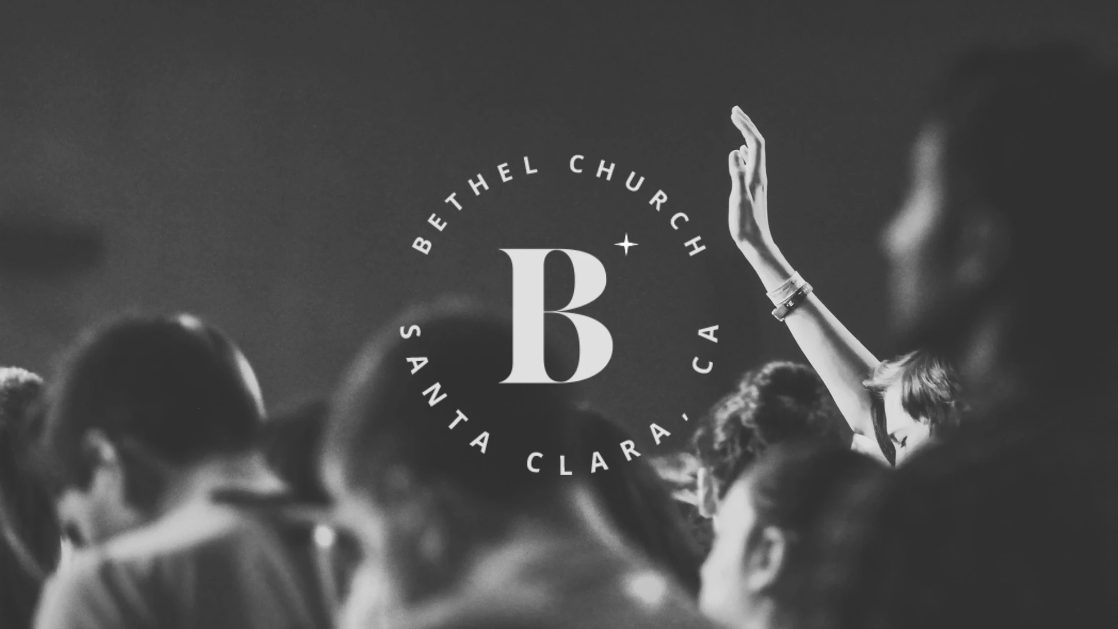 A black and white photo of a church service. One person's hand is raised in worship among a crowd. The Bethel Church Santa Clara logo is overlaid on the image.