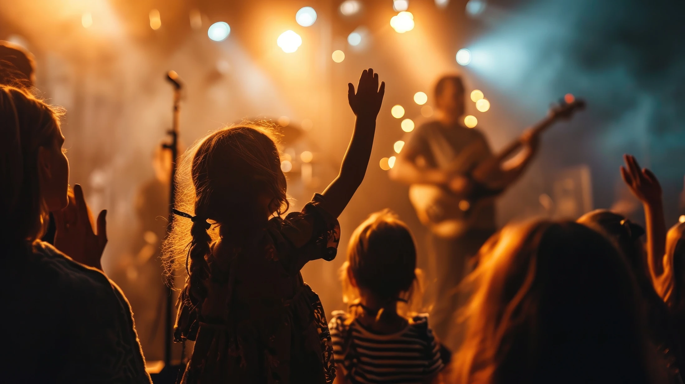 A young girl with pigtails, seen from behind, raises her hand in a crowd at a concert. The stage is warmly lit with a musician blurred in the background.