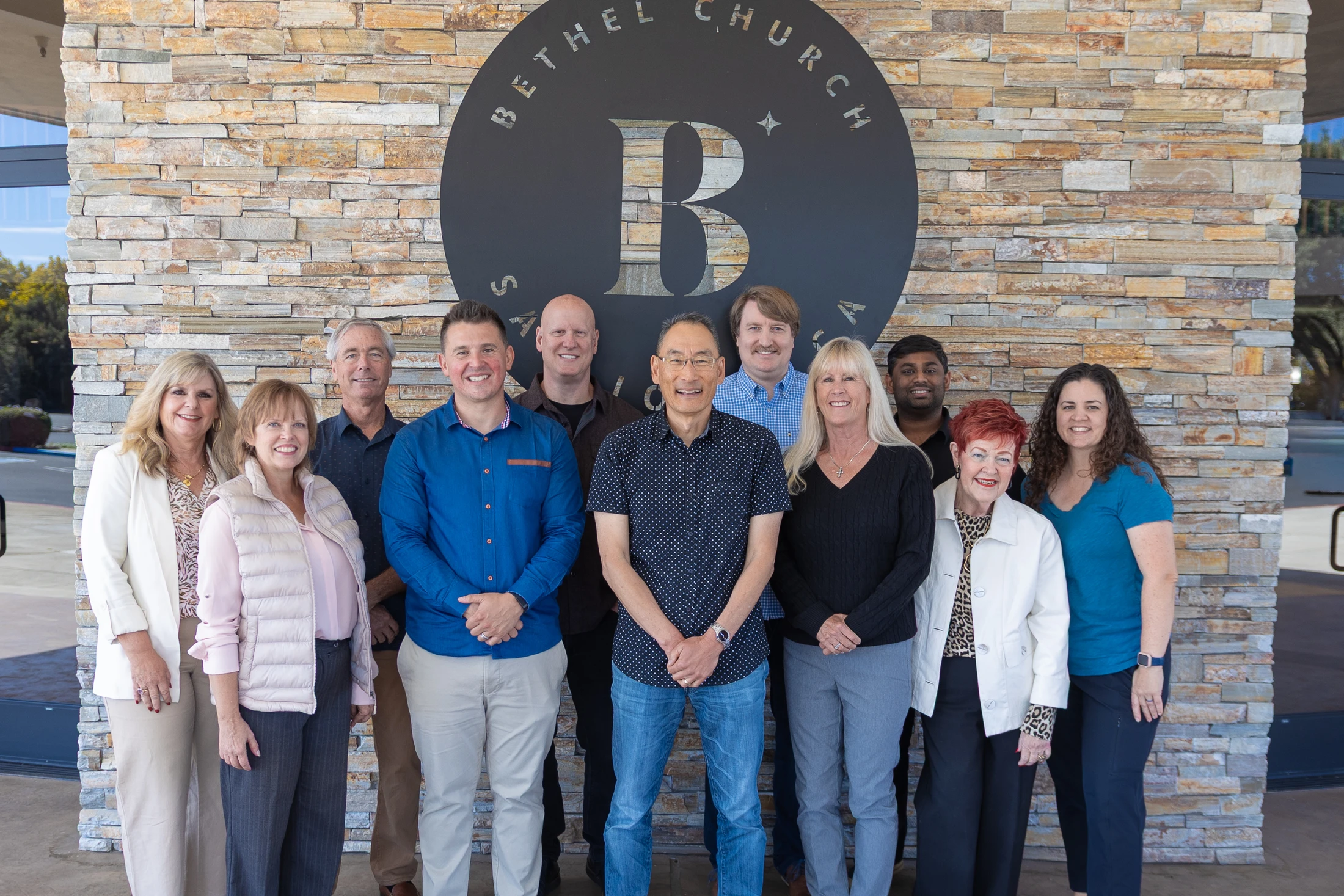A group of eleven smiling men and women pose for a photo in front of a stacked stone wall with a large circular 'Bethel Church' sign.