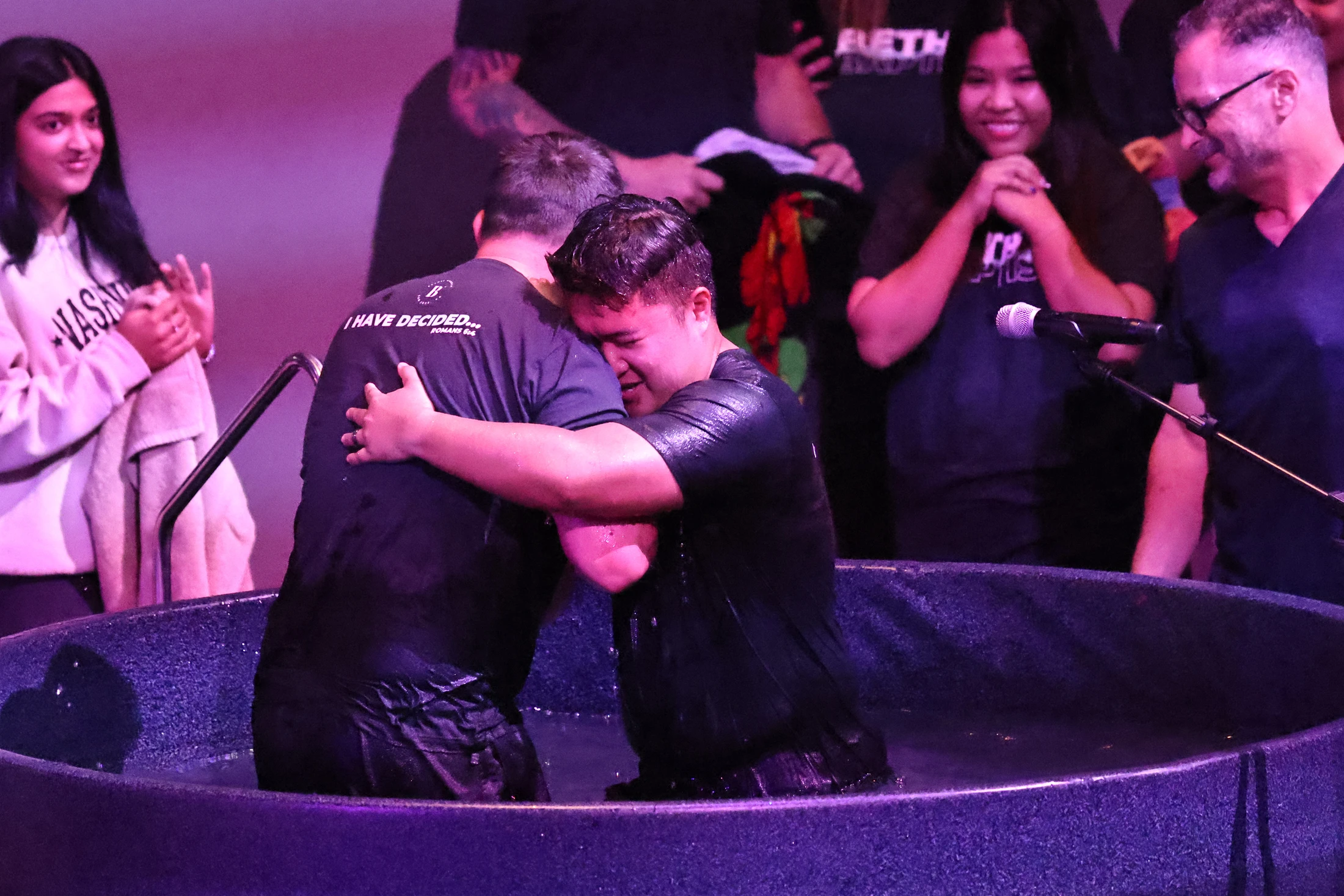 A man cries while hugging another man in a baptismal pool. Onlookers smile in the background during the religious ceremony under purple lighting.