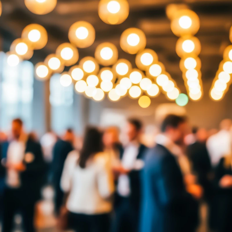 An intentionally blurred image of a corporate event. A crowd of people mingles in a large room under many warm, glowing ceiling lights creating a bokeh effect.