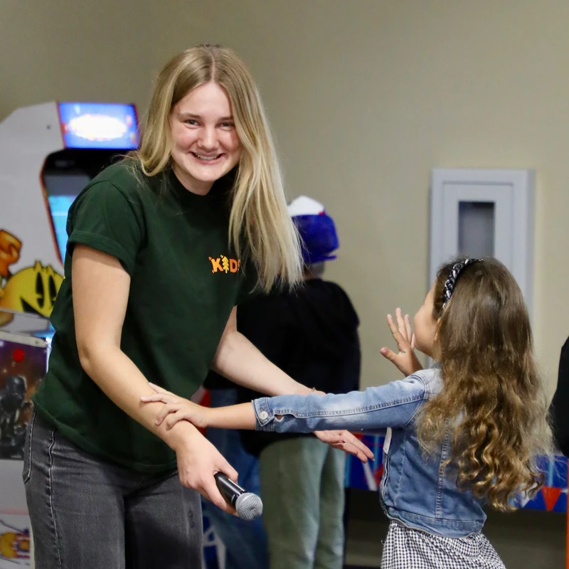 A smiling blonde woman in a green shirt holds a microphone and the hands of a young girl in a denim jacket. An arcade game is in the background.