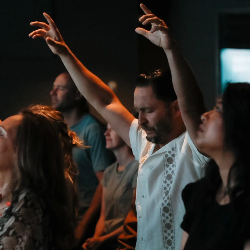 A man with his eyes closed and hands raised stands among a crowd in a dimly lit room, appearing to be worshiping or deeply moved by an event.