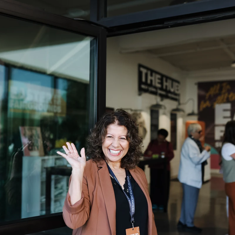 A smiling woman with curly brown hair waves from a doorway. She wears a brown blazer and a lanyard with a 'Here to Serve Dream Team' name tag.