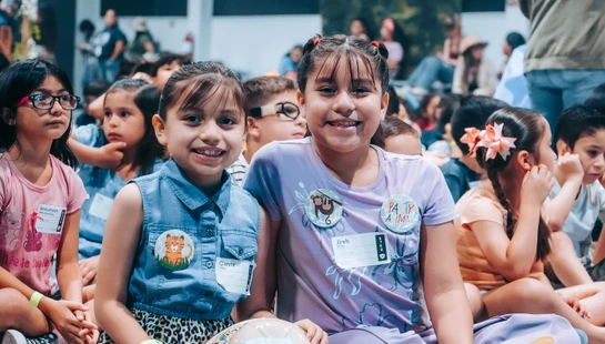 Two young girls smile for the camera while sitting cross-legged on the floor with a large group of other children at an indoor event or assembly.