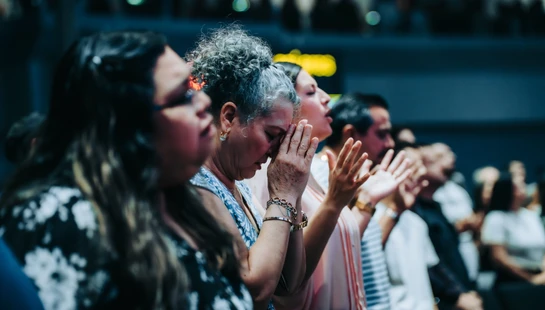 A diverse group of people standing in a row with their eyes closed. The focus is on a woman with gray hair, her hands together in a prayerful gesture.