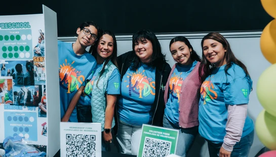 A group of five volunteers in matching blue shirts smile for a photo behind a sign-up table with QR codes and a display board at a kids' event.