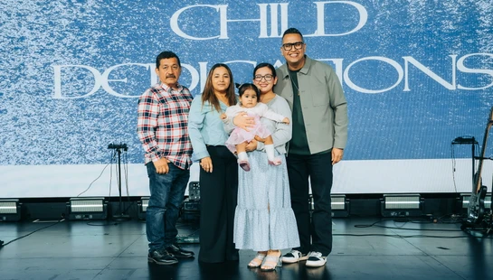 A smiling family of five, including a baby girl, poses on a stage in front of a blue screen that reads 'Child Dedications' during a ceremony.