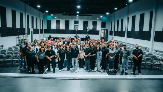 A wide-angle shot of a large, diverse group of people smiling and posing together on a stage inside a modern gymnasium with basketball hoops in the background.