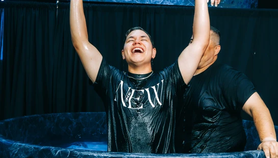 A joyful young man raises his arms in celebration after being baptized in a blue marbled font. He wears a wet black t-shirt that says 'MADE NEW'.