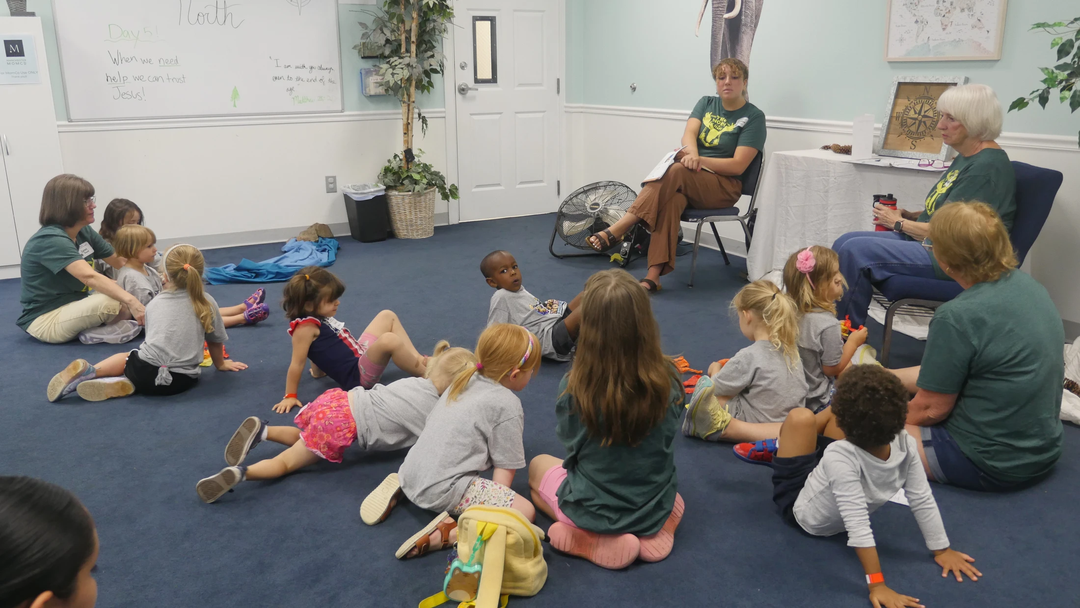 A group of young children sit on a blue carpeted floor, listening to several adult women during a lesson in a classroom setting.