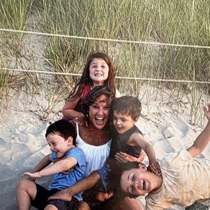 A happy woman sits on a sandy beach, laughing as four young children playfully climb all over her in a joyful pile.