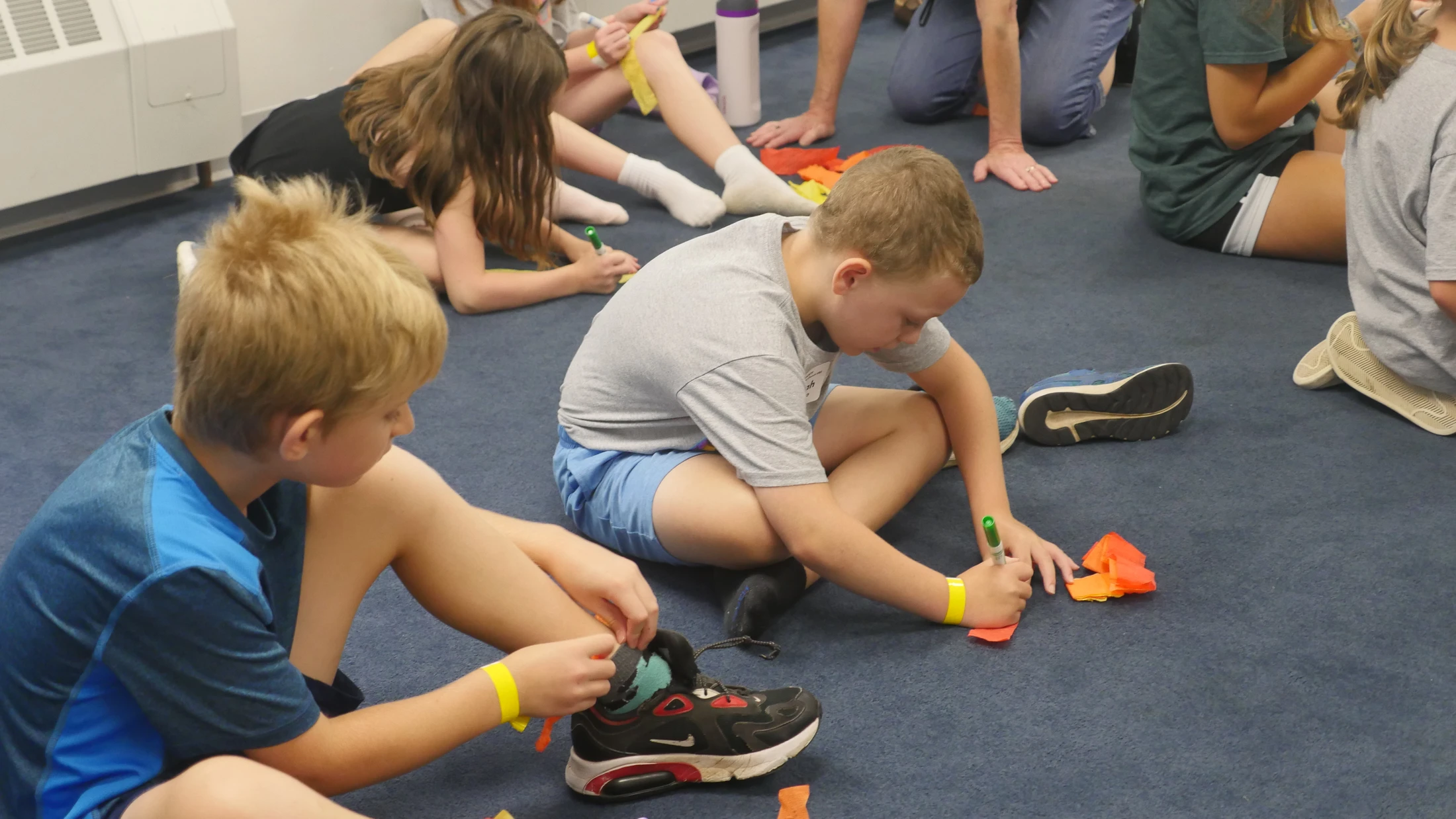 A group of children sit on a blue carpeted floor, focused on an art project with colorful paper and markers, with two boys in the foreground.