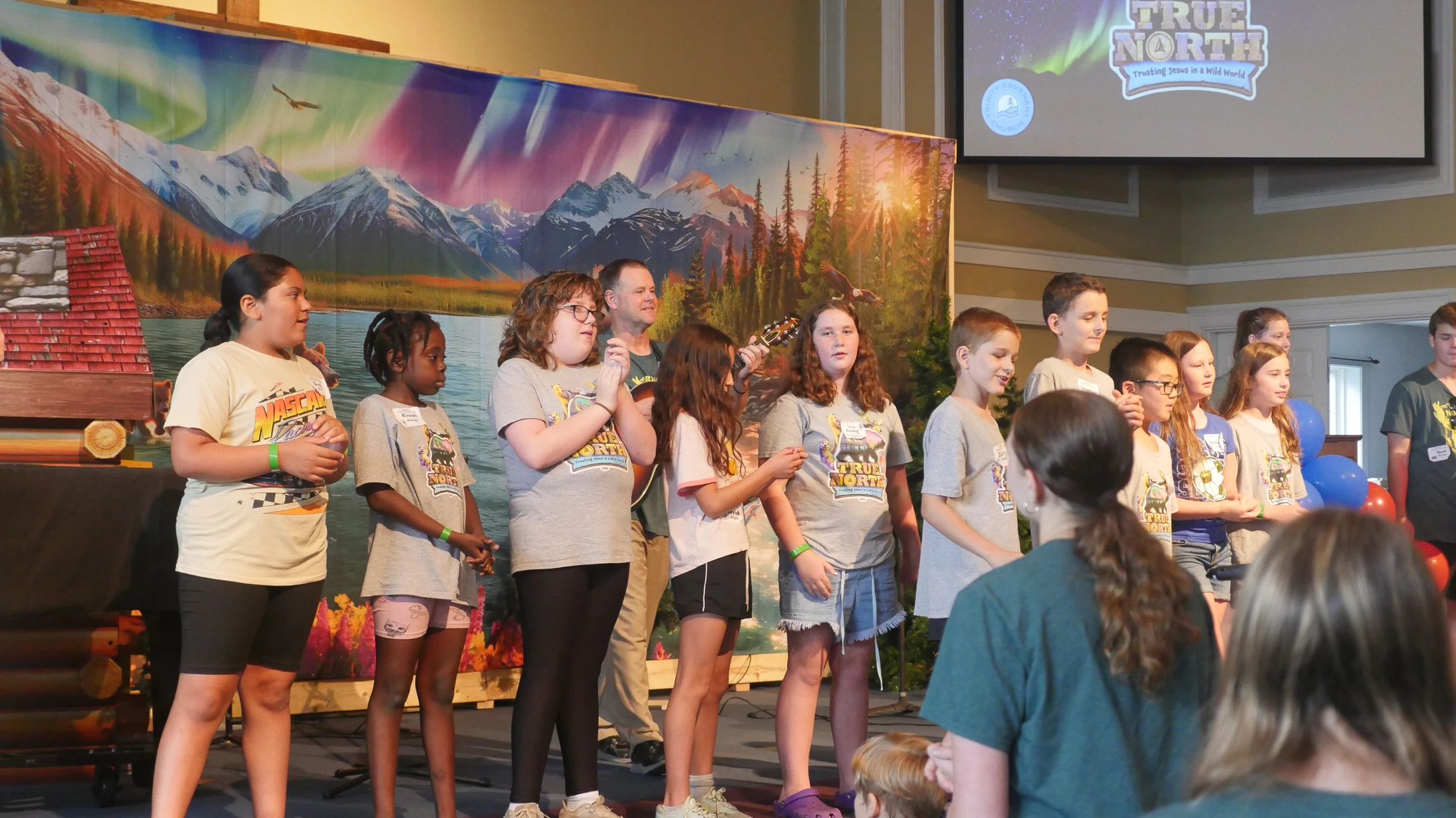A diverse group of children and adults sing on stage in front of a mountain backdrop during a church event called True North.