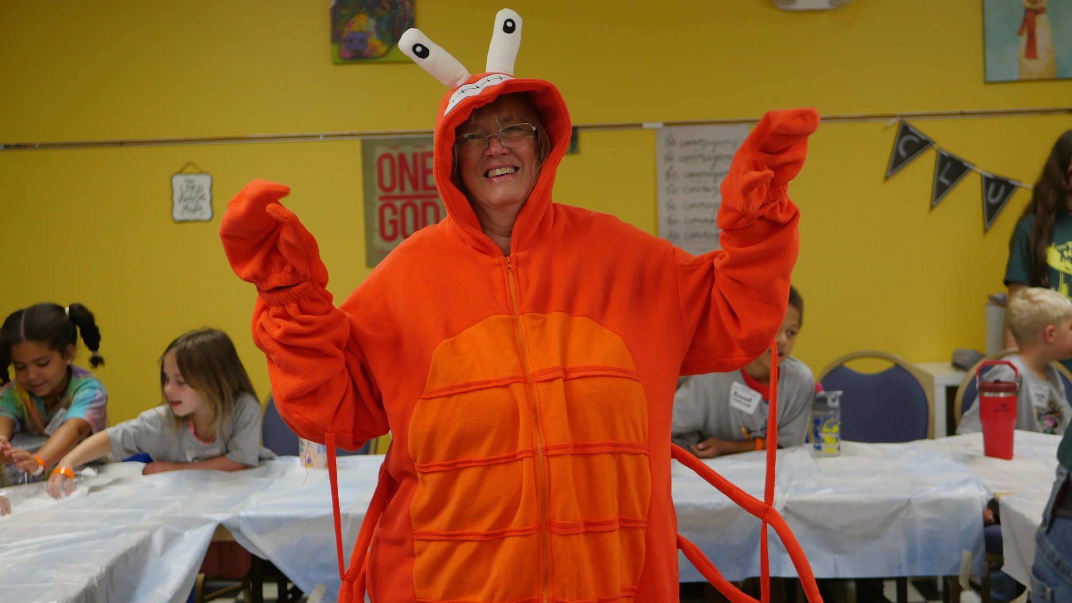 A smiling woman wears a bright orange, full-body lobster costume with big eyes on the hood while standing in a classroom with children in the background.