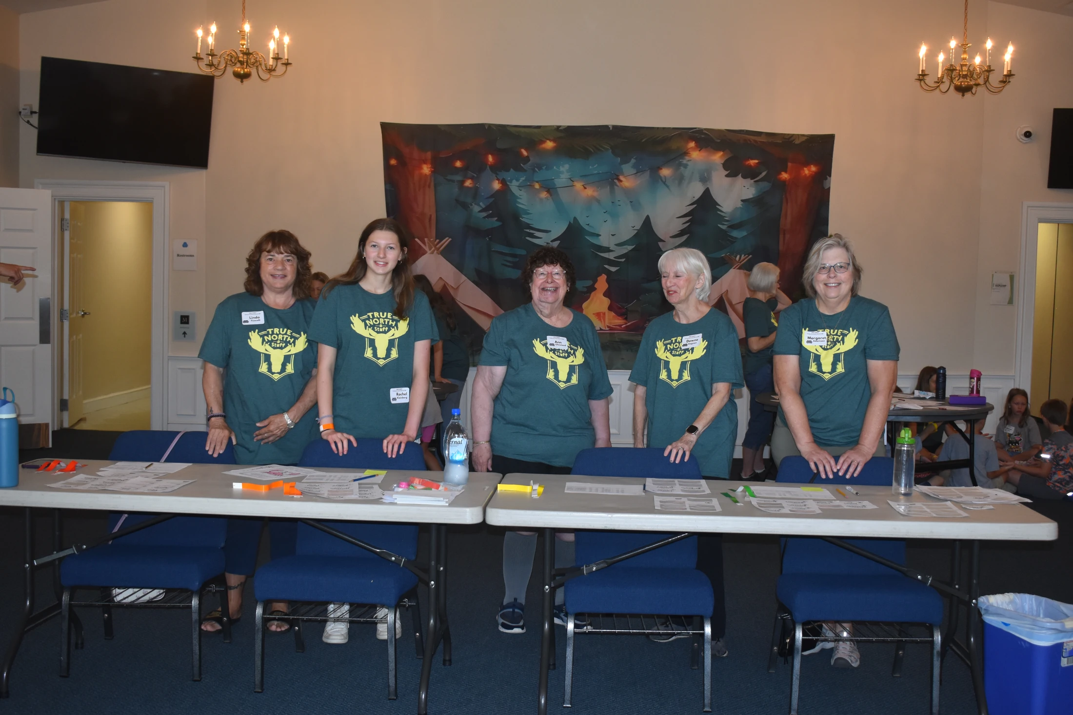 Five women in matching green 'True North Staff' t-shirts smile as they stand behind a long registration table at an indoor event with a forest backdrop.