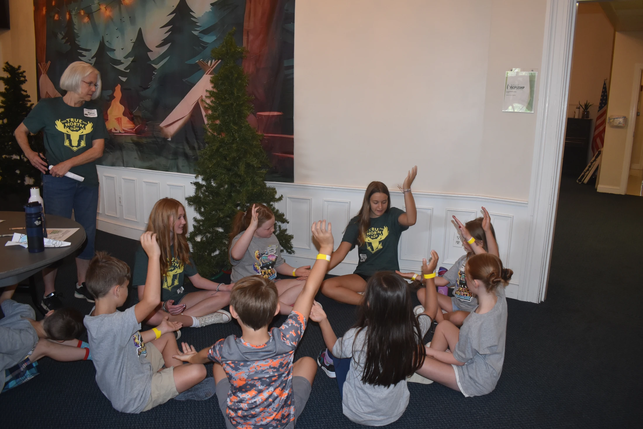 A group of children and a young leader sit in a circle on the floor, playing a game with their hands raised. An adult staff member observes them.