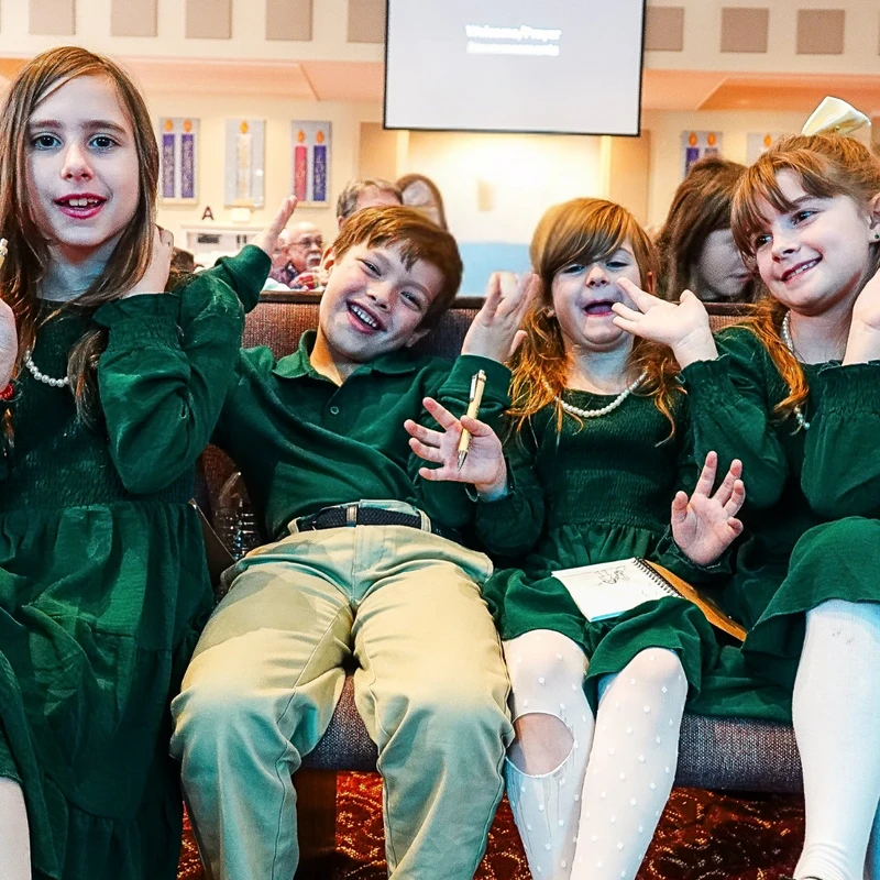 Four children in matching green outfits sit in a row, smiling and making playful faces for the camera while at an event, possibly in a church.