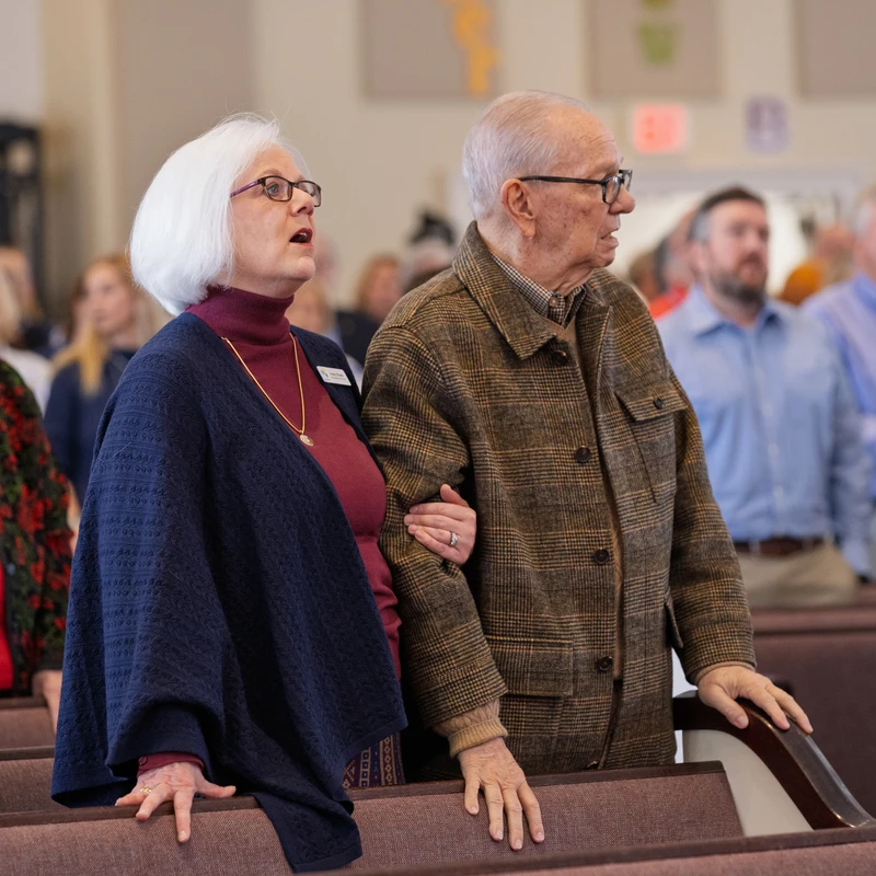 An older couple stands in a church pew. The woman, with white hair and glasses, sings with her mouth open while holding the man's arm.