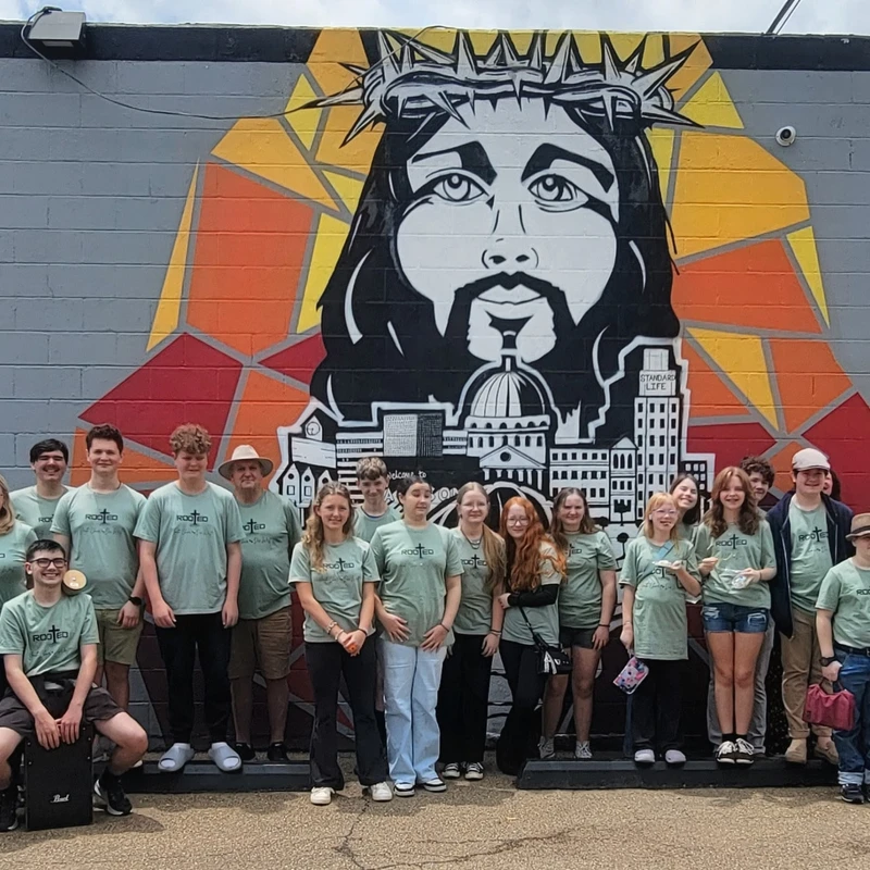A group of teens and adults in matching green 'Rooted' t-shirts stand together smiling in front of a large, colorful mural of Jesus Christ on a brick wall.