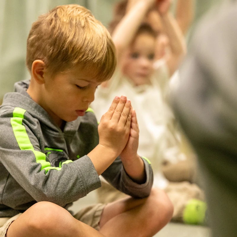 A young blond boy sits on the floor with his eyes closed and hands together in a prayer or meditation pose, looking peaceful and focused.