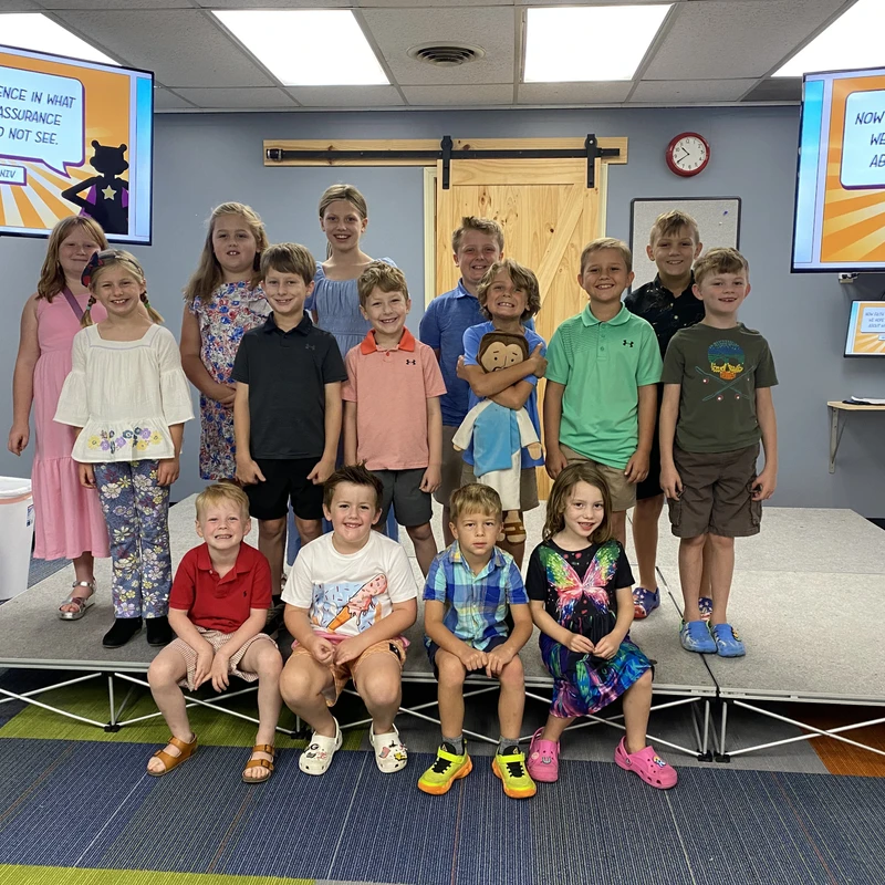 A group of smiling elementary-aged children pose for a photo on a small stage in a classroom, with screens displaying a Bible verse about faith behind them.