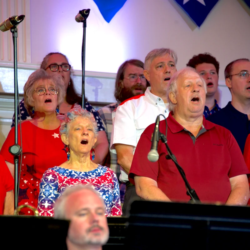 A diverse community choir, dressed in red, white, and blue, sings passionately into microphones during a patriotic performance.