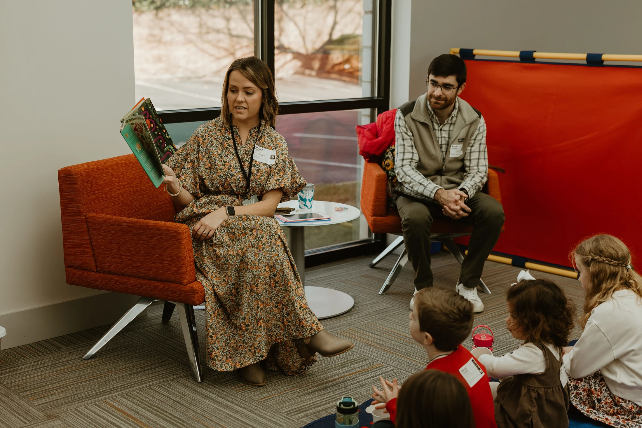 A woman in a floral dress sits in an orange chair, reading a book aloud to a small group of children sitting on the floor in front of her.