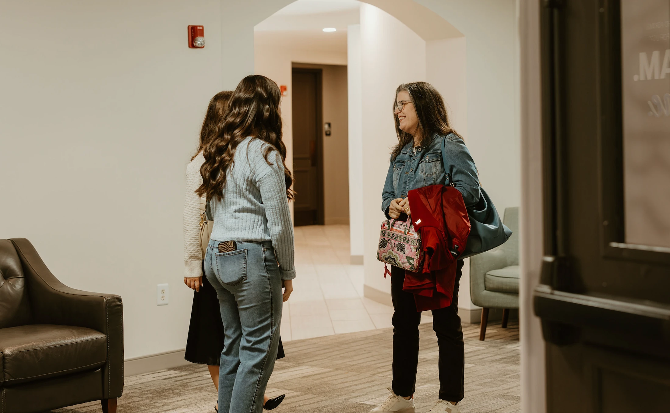 Three women stand in a hallway having a conversation. An older woman in a denim jacket smiles at two younger women who have their backs to the camera.